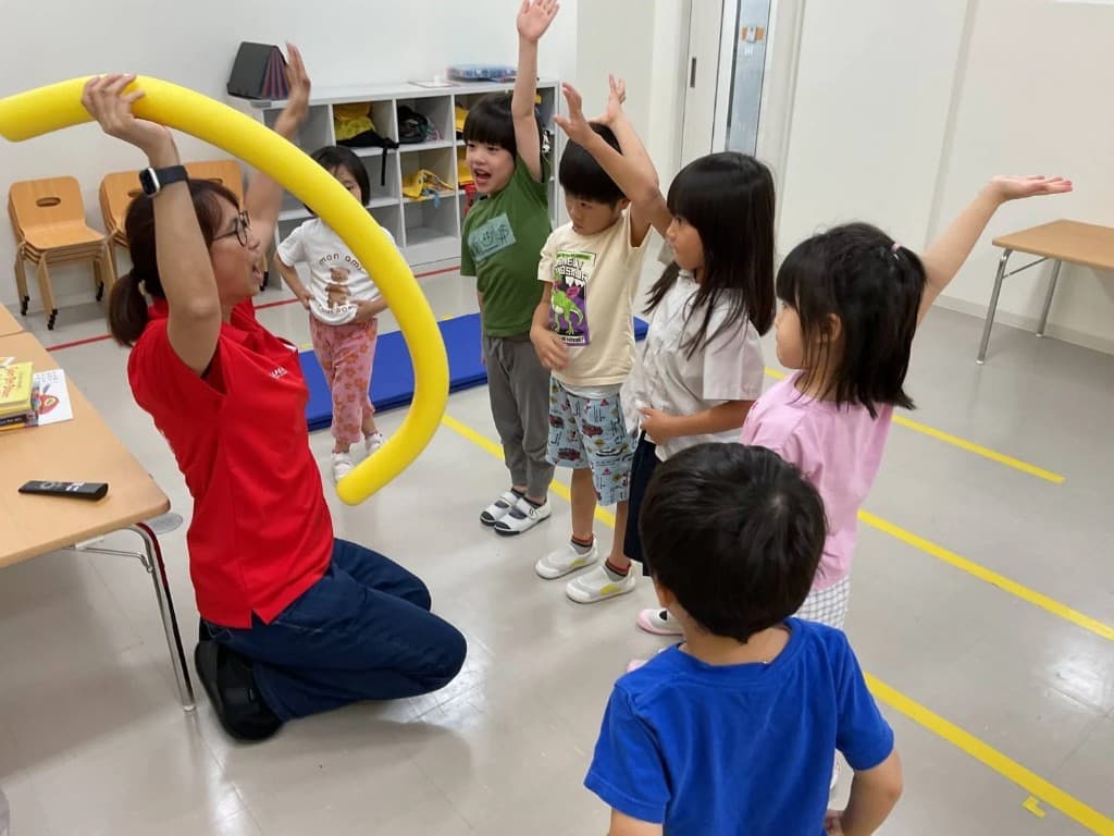 黄色いプールヌードルを掲げ、手を挙げる子どもたちに向き合う講師